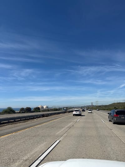 A highway scene under a clear, blue sky with wispy clouds. Multiple cars are traveling in the same direction. The road is gray with painted white lines. The bay is visible beyond a line of trees and a building with a dome-like structure. Power lines stretch across the horizon.
