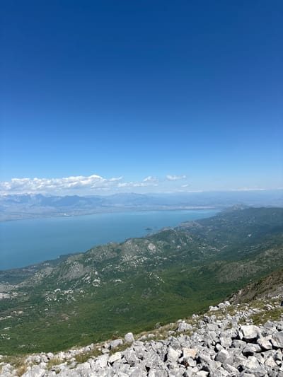 The image shows a view from a rocky mountain edge overlooking a long, narrow body of water, likely a bay or fjord, with green, tree-covered slopes rising on the opposite side. The sky is a clear, bright blue, with a line of clouds along the horizon and at left of the image. The rocky edge in the foreground is composed of large, light-colored rocks. The location is Mali Mikulići, Montenegro.