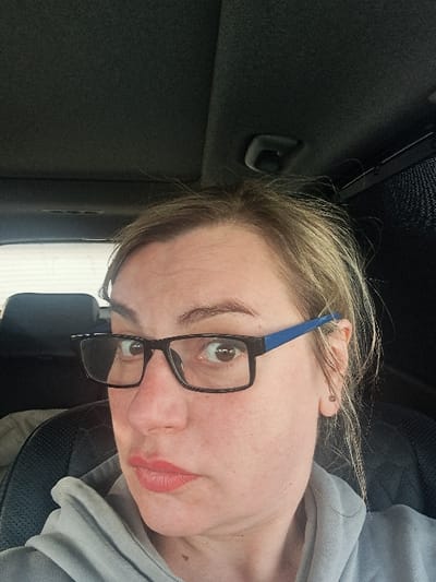 A female subject is depicted in the interior of a vehicle, located in Турлатово, Russia. The subject has light brown hair and is wearing black-framed eyeglasses with blue temples. She has red lipstick applied and is looking directly at the camera with brown eyes. A small, metallic earring is visible in her right ear. She is dressed in a light grey hooded garment. The immediate environment includes a dark grey vehicle headliner, a visible grab handle on the ceiling to the upper right, a dark headrest and textured seat material behind the subject, and a partially visible mesh sunshade on the right side. The scene is illuminated by ambient light.