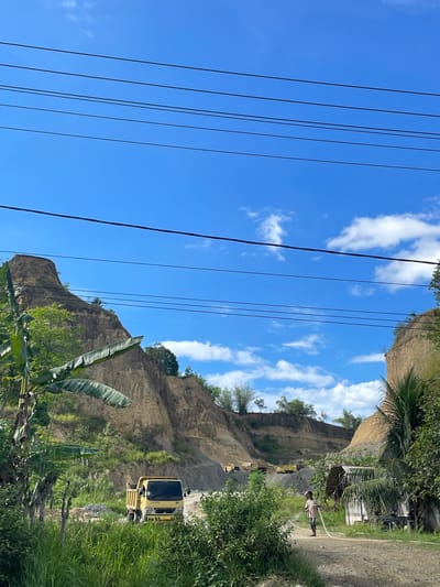 The image depicts an outdoor scene with a bright blue sky and scattered clouds. In the foreground, there is lush green vegetation, including a banana tree on the left. A yellow dump truck is visible on a dirt road, partially obscured by the foliage. Further back, several other yellow dump trucks are parked in what appears to be a quarry or construction site, with large, excavated hills of earth. In the middle ground, a person is standing near a small, simple structure, holding a white hose that is spraying water. The overall environment suggests a rural or semi-rural area, possibly undergoing development or resource extraction. The location is specified as Glee Gogo, Indonesia.