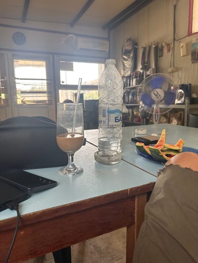 The image shows a still life on a light blue table. The subjects include a nearly empty plastic bottle of water, a glass with a light brown liquid and a straw, a plate with several pieces of watermelon rind, and a person's legs and arm clad in grey fabric. The scene is indoors, with a fan and shelves full of various items visible in the background, suggesting a home or workshop environment. The lighting suggests it is daytime, with natural light coming in from windows in the background, which are partially covered by blinds. There is a clock on the wall showing approximately 11:15. There are also two dark-colored mobile phones, one of which is plugged into a charging cable. The overall impression is one of a casual, perhaps break time moment.
Visible text on the water bottle includes Cyrillic lettering, with "БАР" prominently displayed. The location context is Iskra, Bulgaria.