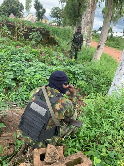 This image depicts two individuals in military camouflage within a densely vegetated, semi-urban environment, located in Sankara, Nigeria. The sky overhead is overcast.
In the foreground, an individual is observed in a crouched position, facing towards the right side of the frame, with their back partially towards the camera. This person wears a dark blue balaclava covering their face, with dark sunglasses positioned over the eye slits. They are dressed in a camouflage military uniform, overlaid with a black tactical plate carrier featuring a light grey rectangular patch on the upper back. A tan strap extends over their left shoulder, likely part of a weapon sling or bag. A weapon, appearing to be a rifle or shotgun with a dark stock, is visible resting along their right side. Their right hand is raised, with fingers near their face, possibly adjusting the balaclava or sunglasses. They are positioned behind a small pile of broken concrete blocks.
In the background, positioned slightly to the right of the frame's center, a second individual stands upright, facing towards the camera and exhibiting a broad smile. This person is also dressed in a camouflage military uniform and wears a tactical vest or chest rig. A rifle, with a dark sling, is visible slung over their left shoulder. They are partially obscured by a light-barked tree trunk to their right.
The environment is characterized by lush green foliage and dense undergrowth covering uneven terrain. Several trees with light-colored, smooth bark are prominent. A dirt path is visible in the distant background, leading towards further vegetation. In the far background, several buildings are visible, including structures with light-colored walls and distinct pointed roofs, suggesting a residential or village area.