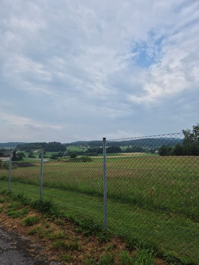 The image depicts a rural landscape in St. Aegidi, Austria, viewed through a chain-link fence. In the foreground, the fence is supported by metal posts, and the ground consists of asphalt and dry grass with some green shoots. Beyond the fence, a field with dry, tall grass and scattered wildflowers stretches towards a rolling, tree-covered hill in the background. Several buildings with red-tiled roofs are visible nestled among the trees and fields, suggesting a small settlement. The sky is overcast with a mix of gray clouds and patches of blue, indicating a partly cloudy day. The overall scene is tranquil and rural.