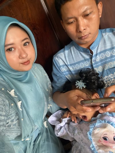An overhead-angle indoor photograph depicts three individuals in Ketegan, Indonesia. In the left foreground, a female with light skin and dark eyes, wearing a light blue hijab and matching dress adorned with white pearl-like beads and white butterfly appliques, looks directly at the camera. To her right and slightly behind, an adult male with light skin and dark hair, wearing a light blue patterned shirt, looks downward. His hands hold a dark-cased smartphone horizontally. Partially visible below the male's hands and to the right of the female is a young child with dark, curly hair secured by a light blue snowflake-shaped hair clip. The child wears a light blue and white patterned outfit, featuring a character image (Elsa from Frozen) on its chest, and has its mouth open. The background consists of a dark wooden surface.