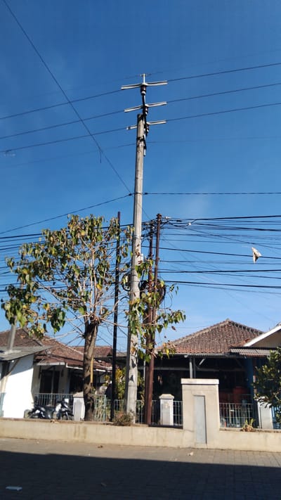 The image depicts a street scene in Bojongloa, Indonesia, under a clear blue sky. The focal point is a tall, concrete utility pole supporting a complex network of overhead electrical wires. These wires extend across the frame, creating a dense web against the sky. To the left of the main utility pole, a smaller, dark pole is visible, also laden with wires. A slender tree with sparse green and brown leaves stands to the left of the main pole, its branches reaching towards the wires.

In the background, several houses with traditional tiled roofs are visible. The houses appear to be single-story structures with white and dark-colored walls. A low concrete wall runs along the front of the properties, separating them from the street. Two motorcycles are parked in front of one of the houses, and a metal fence encloses the property. The street in the foreground is paved with interlocking bricks, casting a shadow from an unseen object to the left. A white plastic bag is caught on one of the overhead wires in the mid-right of the image. The overall impression is of a typical residential street in a warm climate, with the infrastructure of electricity distribution being a prominent feature.