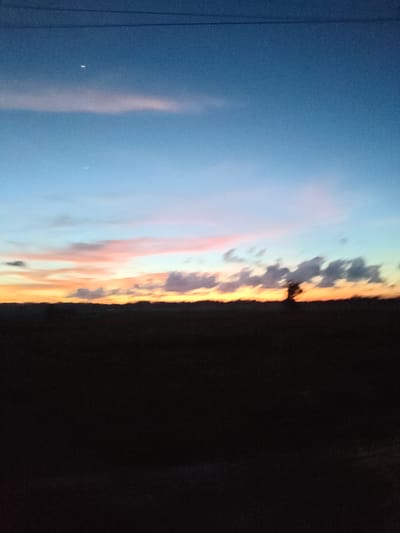 A serene evening scene unfolds in Muliengmanyang, Indonesia, where the sky transitions from a deep twilight blue to hues of orange and pink. Two crescent moons are faintly visible in the upper portion of the sky. A thin, horizontal line, likely a power line, stretches across the top of the frame. Below the sky, a dark, silhouetted landscape stretches across the horizon. A solitary tree stands out against the vibrant sunset, its form a stark contrast to the soft colors of the sky. The foreground is a mass of dark, indistinct shapes, suggesting either vegetation or a field. The overall impression is one of peace and natural beauty as day gives way to night.