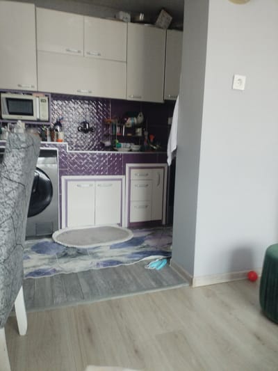 The image depicts a corner of a kitchen area. In the foreground, a portion of a gray and white patterned chair is visible, suggesting it is positioned at a table or counter. The floor is covered with light-colored laminate flooring. Further into the scene, a washing machine is partially visible on the left. Adjacent to it are kitchen cabinets with white doors and light-colored countertops. Above the countertop, there are purple tiled backsplash and upper cabinets. A microwave is mounted above the countertop to the left. On the right side of the image, a light gray wall with a white electrical outlet is present, and a dark green ottoman or pouf is visible in the far right corner. On the floor, near the wall, a small red ball is noticeable. A blue face mask lies on the floor in front of the cabinets. The overall scene appears to be an indoor domestic setting, possibly a kitchen or utility area. There is no indication or context within the image to associate this scene with the city of Rakitovo, Bulgaria.