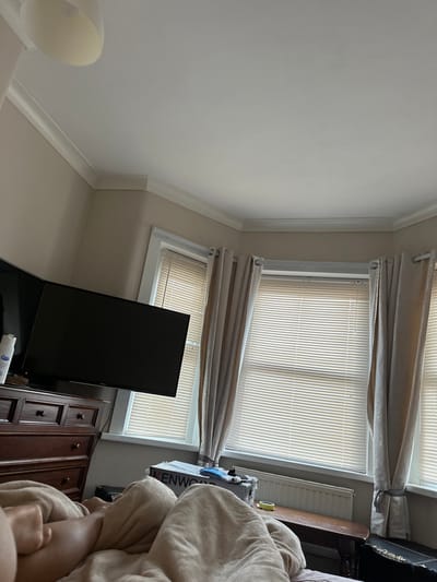 The image displays an interior room located in Dorchester, United Kingdom, viewed from a low perspective. The scene is dominated by a bay window configuration comprising three separate window units. Each window is fitted with light-colored horizontal Venetian blinds, partially closed, allowing filtered natural light to enter. Light beige curtains are drawn back from the windows and secured with decorative ties. To the left of the bay window, a large, black, flat-screen television is mounted on a light beige wall, positioned above a dark wooden dresser with multiple drawers. A white bottle and a small dark object rest on the dresser surface. In the foreground, a person's lower legs are visible, covered by a light beige blanket or duvet. Below the central bay window, a white radiator is present, situated behind a dark wooden bench. A white cardboard box, labeled "KENWOOD," rests on or near this bench, containing what appears to be a blue-handled tool and yellow adhesive tape. A dark-colored trunk or storage box is partially visible in the bottom right corner. The room features white crown molding along the perimeter of the white ceiling, and a white, semi-flush mount light fixture is visible on the upper left ceiling.