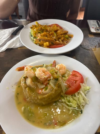 The image depicts an indoor dining scene, likely within a restaurant in San Juan, United States, given the Puerto Rican cuisine. Two white, circular plates containing prepared meals are positioned on a dark, textured table surface.
In the foreground, a plate holds a portion of Mofongo (mashed plantains) partially submerged in a light-colored, thin sauce containing visible pieces of green onion and white onion. Approximately six cooked shrimp are arranged atop the mofongo. To the right of the mofongo, shredded green lettuce and two slices of red tomato serve as a garnish.
In the background, a second white plate displays a dish featuring cooked, flaked fish, consistent with bacalao, mixed with green and red bell peppers, onions, and possibly plantain or root vegetable pieces, all coated in an orange-red sauce. Two red tomato slices are also present on this plate.
A partially visible individual, wearing a brown top, is seated at the table behind the background plate. To the left of the background plate, a silver-bladed knife with a black handle rests on a crumpled white napkin. To the right, a mobile phone encased in a light-colored, patterned cover with a circular accessory is visible on the table, which exhibits water marks. The scene suggests an active dining environment.