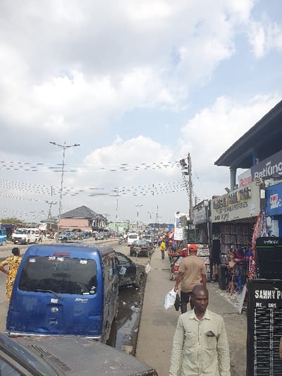 The scene depicts a bustling urban street in Igwuruta, Nigeria, under a partially cloudy sky. In the foreground, the hood of a dark vehicle is visible at the bottom. A blue Daihatsu Atrai van, marked "TOPMAX," is prominently parked, angled away from the viewer, with its rear left wheel positioned in a roadside ditch containing standing water and debris. To its immediate right, a dark sedan appears partially submerged in the same ditch, showing significant damage to its driver's side door area.
On the sidewalk in the foreground, a man in a light green long-sleeved shirt walks towards the camera, looking straight ahead. To his right, a tall, black display stand with an "Infinix" logo at the top and "SAMMY P" and "M/C DRG" visible below shows a grid of information. Further down the sidewalk, another man wearing a tan shirt and a red cap walks away from the camera, carrying a white plastic bag.
The right side of the street features multiple commercial storefronts, with signs such as "MARISON PERFUMES" and "BetKing." Merchandise, including wigs on mannequins, is displayed outside these establishments. The paved road extends into the distance, populated by various vehicles including cars, vans, and motorcycles, indicating active traffic. Numerous utility poles with complex networks of overhead wires and multi-arm streetlights line the street. Buildings of varying architecture extend along both sides of the thoroughfare, receding into the background. A woman in a patterned yellow dress is visible on the far left sidewalk, looking towards the road.