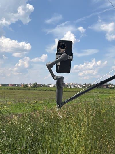 The image shows a smartphone mounted on a gimbal, held by a black pole. The smartphone is black and has a prominent camera array on the back. The gimbal is also black and has a white string attached to it. The scene is outdoors, with a bright blue sky filled with white, fluffy clouds. A green field stretches into the background, with a line of houses visible in the distance. The overall impression is one of a clear, sunny day. The image is oriented vertically, suggesting it might have been taken with a handheld device. There is no activity or event clearly depicted, other than the setup of the filming equipment. The location is specified as Klisheva, Russia, which aligns with the rural scenery. The time of day appears to be daytime, likely late morning or afternoon due to the bright sunlight. No people are visible in the image. Text visible on the smartphone reads "HUAWEI".