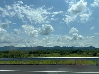 A wide-angle, eye-level shot captures a roadside view with a mountainous, forested landscape under a partly cloudy sky. The foreground features an asphalt road with a white stripe, bordered by a metal guardrail and grassy verges. Beyond the guardrail, a field of green grass stretches towards a dense line of trees. Further in the distance, rolling hills covered in lush green vegetation form a backdrop, culminating in the silhouette of mountains against a bright blue sky dotted with fluffy white clouds. The scene appears to be captured from a vehicle moving along a road in the rural area of Chibat'is Temi, Georgia. There are no people or animals visible in the frame.