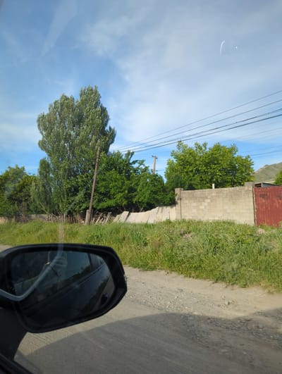 The view from a moving vehicle shows a roadside in Beş-Küŋgöy, Kyrgyzstan. The sky is partly cloudy with power lines running overhead. Green trees line the road, next to a concrete block wall topped with a red roof. A dirt road runs alongside the green vegetation. The car's side mirror is visible in the foreground.
