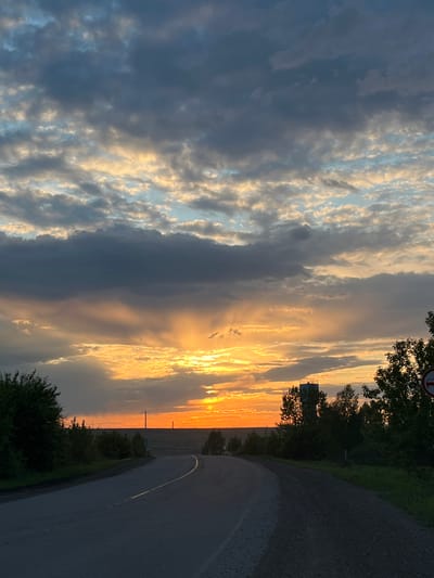 The image depicts a road in Zykovo, Russia, under a sky with dramatic clouds at sunset. The road curves into the distance on the right side of the frame. Along the right side of the road are trees and bushes. The sky features a mix of dark, heavy clouds and bright, golden clouds illuminated by the setting sun. The sun's rays filter through the clouds, creating a striking contrast. A road sign is visible among the trees in the upper right corner. A utility pole is visible near the horizon. There are no people visible.