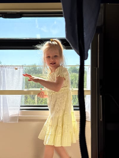 A young Caucasian girl with blonde hair tied in a ponytail, wearing a light yellow dress, is standing by a window with white curtains. She is looking at the camera with a slight smile and has her hands extended. Outside the window, trees are visible against a blue sky. The scene appears to be indoors, possibly in a train or bus, given the window structure and a dark curtain on the right side of the image. The location in Grushevskaya, Russia is not discernable from the image itself.