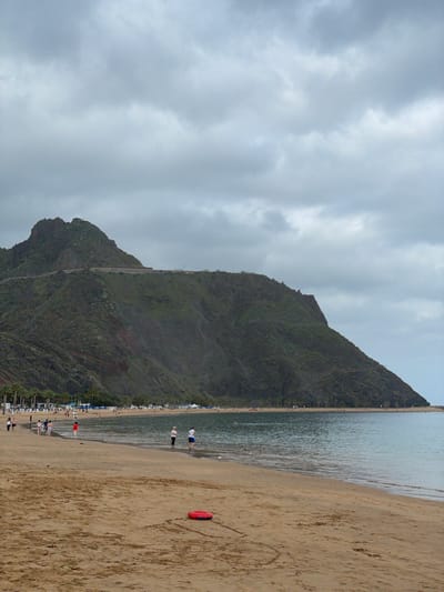 The image displays a serene coastal landscape, likely situated in Santa Cruz de Tenerife, Spain, under an overcast sky. The scene is dominated by a large, dark green, vegetated mountain that rises steeply on the left side of the frame, directly from the coastline. A faint, lighter-colored path can be seen winding its way up the mountain's face.
To the right of the mountain, a long, sandy beach stretches into the distance, meeting the calm, blue-green ocean water. The water shows gentle ripples and appears tranquil. Several people are visible along the shoreline; two individuals are seen wading in the shallow water near the center, and others are scattered further down the beach. In the background, along the base of the mountain, rows of beach chairs and blue umbrellas suggest a leisure or resort setting.
A distinctive red, U-shaped inflatable ring or buoy lies on the sand in the mid-right foreground. The sky above is entirely covered by soft, grey clouds, indicating an overcast day, which casts a diffused light over the scene. The overall activity appears to be one of relaxation and leisure by the sea. No visible text is identifiable within the image.