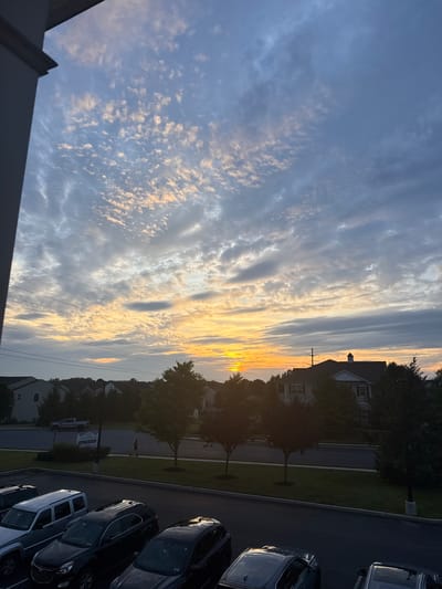 The image captures a scenic view of a sunset over Allen Township, United States of America. The sky is adorned with a mix of cirrus and cumulus clouds, painted in hues of orange, yellow, and blue, suggesting the sun is near the horizon. Silhouetted against the vibrant sky are several residential buildings and lush green trees. In the foreground, a parking lot is visible, occupied by several parked cars, including silver, black, and gray SUVs. A person is seen walking on a sidewalk in the middle ground, appearing to be enjoying the evening. The overall atmosphere is serene and peaceful, with the fading daylight casting a warm glow on the landscape.