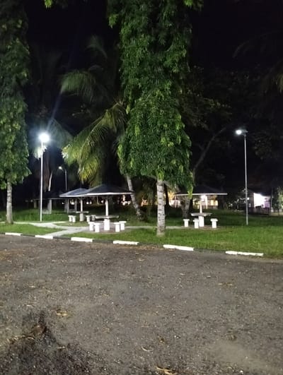 This media file depicts a nighttime scene at Palohgadeng, Indonesia. The setting appears to be a park or recreational area, illuminated by artificial lighting. In the foreground, a paved surface, possibly a road or parking area, is visible, covered with scattered leaves and debris.
The mid-ground features several covered picnic shelters with attached concrete tables and benches. These shelters have dark, angular roofs and are positioned on a patch of green grass. Two prominent lampposts with bright lights stand in the grassy area, casting a glow on the surroundings. Further in the background, more trees, including palm trees, are visible against the dark night sky. Another structure, possibly a building or a larger shelter, is dimly lit in the far background.
The time of day is clearly night due to the darkness and artificial lighting. The weather appears to be clear. No specific activity or event is taking place; the scene is static and empty. Notable details include the presence of mature trees, the design of the picnic shelters, and the lighting fixtures. The overall mood is calm and perhaps a little deserted due to the absence of people and the nighttime setting. There is no visible text in the image.