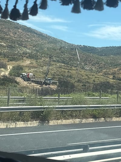 This image depicts a construction scene on a hillside in Avra, Greece. The primary subjects are two construction vehicles, likely a concrete mixer and a pump truck, positioned on a dirt road amidst scrubby vegetation. A person wearing a white shirt and what appears to be a red and white striped top is visible near the vehicles, possibly overseeing the operation or working. The vehicles are angled on the slope, suggesting they are engaged in pouring concrete.
In the background, a steep, vegetated hillside rises towards a bright blue sky with wispy white clouds. A fence line with vertical posts runs along the right side of the scene, separating the construction area from what appears to be a paved road with distinct white lane markings. The light suggests it is daytime, likely late morning or early afternoon, with clear weather. The overall impression is one of ongoing civil engineering work in a natural, hilly landscape. There is no visible text in the image.