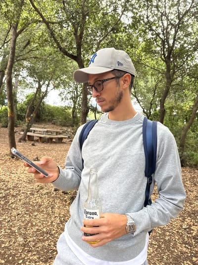 A man stands in a wooded area, likely a park, location unspecified, although presumed to be near Nesher, Israel. He is wearing a grey sweatshirt, grey baseball cap with a blue logo, glasses, and a silver watch. He is wearing a backpack. He holds a cell phone in his left hand and a bottle of Corona Extra beer in his right. Fallen leaves cover the ground. A picnic table is visible in the background. The lighting suggests a sunny day.