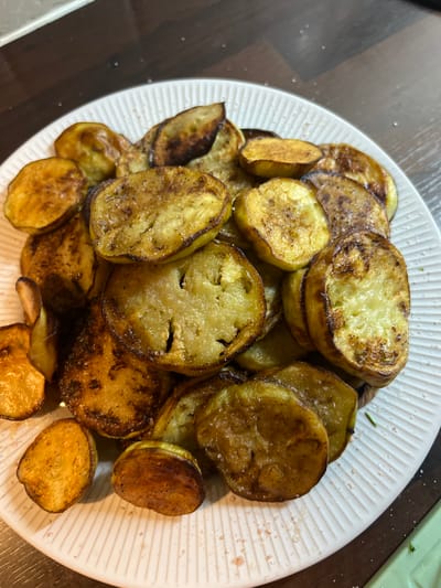 A close-up, overhead shot of a white, ribbed plate piled high with fried eggplant slices. The eggplant is golden brown with some darker, caramelized edges, and is lightly sprinkled with salt. The plate sits on a dark wooden surface. There are no people visible in the image, and no specific location in Balbriggan, Ireland can be determined from the provided visual information.