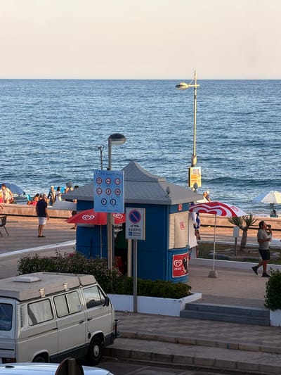 The image depicts a scene along a seaside promenade in Castillo Bajo-conejito, Spain. The sky is a pale, cloudless hue, transitioning to a calm, blue ocean. A paved promenade runs parallel to the sea, with a white-tiled wall separating it from a grassy area.
On the promenade, several people are visible. Some are seated on beach chairs or towels near the water's edge, partially obscured by blue umbrellas. Others stand or walk along the promenade. A man in a dark blue shirt and white shorts stands facing the sea, while another man in a grey t-shirt and dark shorts walks away from the camera, holding a water bottle.
A prominent blue kiosk with a grey roof is situated on the promenade. It has signs and awnings. One sign, a large blue rectangular placard, displays multiple circular icons with red borders, indicating regulations for the promenade. Another smaller sign below it shows a red circle with a diagonal line over a wheelchair symbol, and text that reads "EXCEPTO MOVILIDAD REDUCIDA." A red and white striped umbrella is positioned in front of the kiosk. Two red Frigo ice cream logos are visible on the kiosk's side.
To the left of the kiosk, a white camper van is parked. It has several windows and a raised roof section. A blue wheelchair symbol is affixed to one of the van's windows.
Further down the promenade, a lamppost stands near the kiosk. A small yellow sign is attached to the lamppost with the text "FOOD TRUCK ESPINOS TORREX." Another, larger white umbrella with a blue canopy is visible in the distance near the water.
The ground is paved with interlocking diamond-shaped tiles. In the foreground, a section of a road or lower pathway is visible with a metal grate. Lush green bushes and small pink flowers line parts of the promenade, particularly near the kiosk and the camper van. The overall impression is a public space by the sea, likely during daytime hours, with people enjoying the coastal environment.