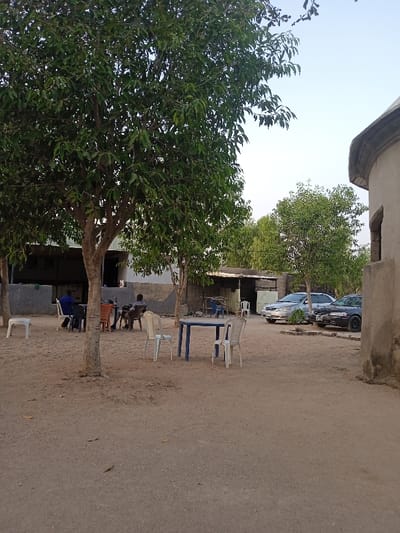 An outdoor scene shows a sandy ground area with trees offering shade. A blue table with white chairs sits in the center. People are seated at tables under the trees. A silver and black car are parked nearby. In the background, there's a building and a small shack-like structure. To the right, a portion of a weathered, dome-shaped building is visible. The sky is a pale blue.