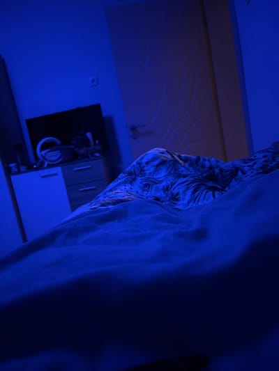 The image depicts an indoor scene bathed in deep blue artificial light, suggesting nighttime or a mood-lit environment. On the right side, a substantial amount of light-colored fabric, possibly a curtain or sheet, drapes downwards with visible folds. Adjacent to this, in the mid-ground, is another piece of fabric, possibly clothing, featuring a distinct pattern of dark, foliage-like or abstract shapes against a lighter background.
The left side of the frame reveals a wall or ceiling, also tinted blue. Towards the lower-left, a dark piece of furniture, possibly a cabinet or dresser, is visible. On its surface, several indistinct objects can be seen, including a white, circular item that resembles a mug or bowl, and other metallic or reflective items. Higher on the wall, a light-colored electrical outlet or switch is present. The upper part of the wall or ceiling shows a subtle decorative pattern, possibly stucco or raised wallpaper. There are no people visible, nor any discernible text. The overall atmosphere is serene and subdued, created by the monochromatic blue illumination. The location context is Nadarevo, Bulgaria.