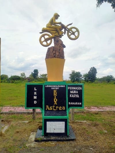 A statue depicts a motorcyclist on a rock formation atop a tall, yellow pedestal. Beneath it, a sign with green trim displays black panels with text: "LKM D" to the left, "LAPANGAN OLAHRAGA SIRCUIT KRIDA ASTREA" in the center, and "PERMASDA MUDA KARYA BANARAN" to the right. Below, a smaller section houses square, white tiles. The statue and sign are situated in a grassy area with trees in the background under a cloudy sky.