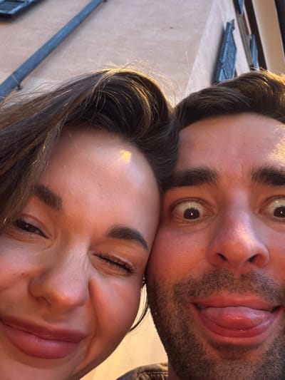 This is a close-up, high-angle selfie taken outdoors in Nobiallo, Italy, showing two young adults. The woman on the left is winking and smiling, with her hair falling across her forehead. The man on the right has his eyes wide open and is sticking out his tongue, with stubble on his chin. The background consists of a textured wall and a dark pipe. The lighting suggests it is late afternoon or early evening.