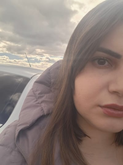 A close-up selfie of a woman with brown hair, light skin, and pink lipstick. She is wearing a light purple jacket. The background shows a car and a cloudy sky. The location is not possible to determine only by this picture, the city of Povarenki in Russia cannot be verified.
