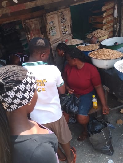 This image captures a vibrant market scene in Mgbuoshimini, Nigeria, under bright daylight. Several people are gathered around a bustling stall selling dried foodstuffs.

In the foreground, the back of a person with braided hair and a black and white patterned headband is visible, wearing a dark top. Next to them, another individual is seen from behind, wearing a white polo shirt with green stripes on the collar and sleeves, and brown shorts. The partial text "MA GOLD CE" is printed on the back of the shirt, possibly part of a longer phrase like "OBAMA GOLD PEACE." This person appears to be observing the goods or interacting with a vendor.

To the right, a woman with dark hair, dressed in a red short-sleeved top and blue denim shorts, is seated on a low wooden stool. She holds a black plastic bag, and a yellow plastic bottle rests beside her on the stool. Another large black bag is visible on the ground near her, along with scattered coconut husks. Further back, another woman with dark hair is partially visible.

The market stall features a display of various dried grains or legumes, likely peanuts, beans, or corn, presented in large white and blue enamel bowls on a counter. The background of the stall consists of old, weathered wooden or metal doors, adorned with several stickers and labels, some partially readable such as "GRACE" and "DIFFERENT." Stacked sacks are also visible behind the counter, indicating more inventory. The overall scene conveys the active, communal atmosphere of a local market.