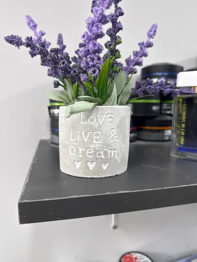 This is a close-up, vertically oriented photograph of a small, grey, concrete pot with the embossed text "LOVE LIVE & dream" and decorative hearts. The pot contains artificial lavender sprigs and green faux leaves. The pot is placed on a dark grey, possibly wooden, surface, which appears to be the edge of a shelf or stand. In the background, several camera lenses and a glass bottle of what appears to be perfume are visible. The lighting suggests an indoor setting, and there are no indications of weather or specific time of day. The overall mood is decorative and still life-like. The location is identified as Gurkovo, Bulgaria.
