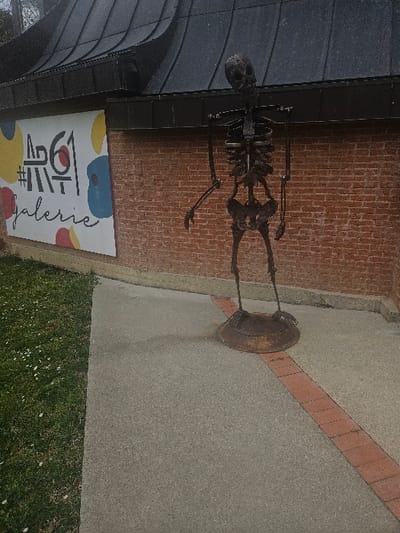 An outdoor scene in Avignonet-Lauragais, France, depicts a dark metallic skeleton sculpture standing on a concrete sidewalk in front of a red brick building. The sculpture, approximately human height, is posed upright with arms slightly bent and away from its torso, and rests on a wide, rusted, circular metal base. To the immediate left of the sculpture, a white rectangular sign with the text "#ART61 galerie" in black and abstract yellow, red, and blue shapes is affixed to the brick wall. Above the wall, a dark, corrugated or paneled metal roof with a distinct curve is visible. The concrete sidewalk extends from the lower left foreground, featuring a diagonal strip of reddish-brown bricks, and meets a patch of green grass dotted with small white flowers on the left.