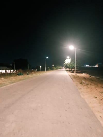 The image shows a street at night, likely in Rafin Pa, Nigeria. The street is illuminated by sparsely spaced streetlights. The road surface appears to be asphalt or concrete. Vegetation lines the sides of the road, with trees near the streetlights. Buildings are visible in the distance, lit by their own lights. The scene is dark, and the focus is on the illuminated road leading into the distance. There are no visible people or vehicles in the immediate view.