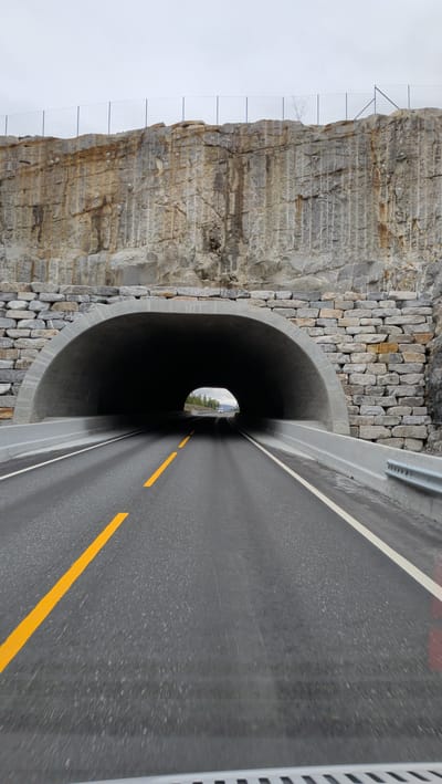 This media file shows a perspective view of a road leading into a tunnel in Grane, Norway. The tunnel entrance is arched and constructed with concrete, with a stone facade on the upper portion of the surrounding rock face. The rock face itself appears to be a quarry, with vertical cut marks and some areas of natural stone. There is a wire fence at the top of the rock face. The road is asphalt and has a yellow dashed line on the left side and a solid white line on the right. A metal guardrail is visible on the right side of the road. The weather appears overcast, and the time of day is unclear. In the distance, through the tunnel, the sky is visible with trees and a white vehicle. The overall scene conveys a sense of a natural landscape modified for infrastructure. There is no visible text in the image.