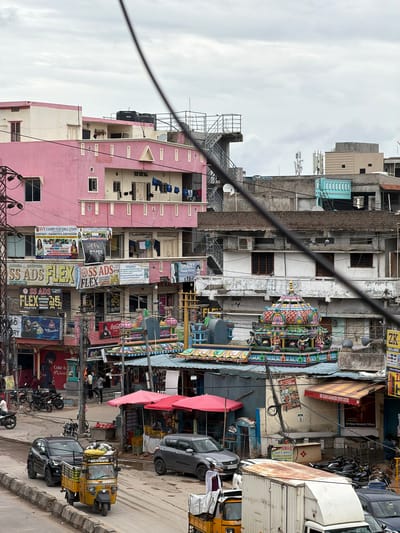 This is an eye-level, medium shot capturing a street scene in Shamshabad, India, under an overcast sky. The foreground is dominated by a yellow auto-rickshaw carrying goods, followed by a black SUV and a silver sedan. Beyond the vehicles, the street is lined with a mix of shops and multi-story buildings.
A prominent pink building on the left features multiple floors with windows and balconies, where laundry is hung out to dry. Various businesses are advertised on banners and signs attached to the buildings, including "SVV COMPUTER EDUCATION," "SAHASRA ADVERTISING," and "SS ADS FLEX." Further into the scene, a vibrant, colorful temple structure with intricate carvings and statues is visible. The temple has a tiered roof, painted in shades of pink, green, yellow, and blue, and is adorned with small figures of deities.
Adjacent to the temple are more shops with awnings, displaying goods for sale. Several motorcycles are parked along the roadside. People are seen walking on the street and in front of the shops. Utility poles and a dense network of electrical wires crisscross the view, indicating a busy urban environment. The buildings display a variety of architectural styles, from simple concrete structures to more ornate designs. The overall impression is of a bustling commercial area with a blend of traditional and modern elements.