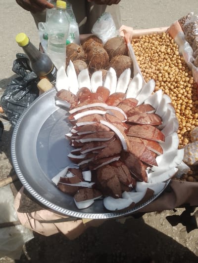 An overhead shot shows a street vendor's cart displaying various food items. A large metal platter is filled with chopped coconut pieces, some with white flesh and others with brown husk. Scattered around the platter are whole coconuts, as well as bags of what appear to be nuts or seeds. A plastic bottle of water and a dark-colored bottle with a yellow cap are also visible. The scene suggests a market or street-side stall in a city like Beli, Nigeria, where such fresh produce and snacks are commonly sold.