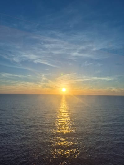 The image shows a horizontal division of sky and sea at sunset. The sun is positioned at the horizon line, emitting a bright golden light that reflects on the water. The sky exhibits varying shades of blue with wispy clouds. A structure with geometric lines is faintly visible in the upper left corner, suggesting a building or infrastructure. The location is Unknown, France.