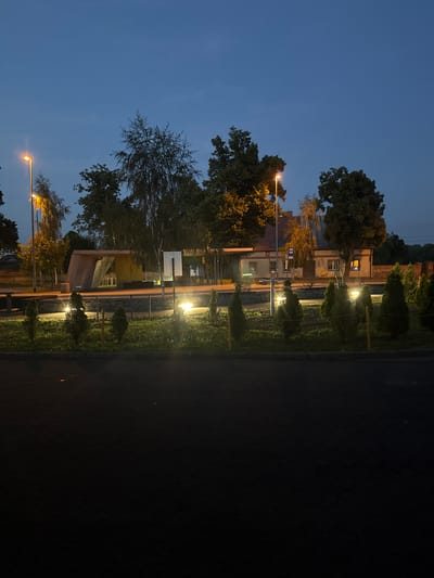This image depicts an outdoor scene at dusk or early night in Wąwelnica, Poland. In the foreground, a dark asphalt or paved area with a visible brick-like pattern stretches across the bottom of the frame. Beyond this, a low-lying grassy area is illuminated by several ground lights, casting a warm glow. Several small, cone-shaped evergreen trees are interspersed within this grassy area, with thin wooden posts marking the boundary.
A low wall made of stone blocks forms a border between the grassy area and a walkway or platform. Behind the wall, a modern-looking structure with a slanted concrete roof and yellow panels is visible, possibly a bus stop shelter or a small pavilion. Further back, a traditional building with a tiled roof, windows with illuminated interiors, and a bus stop sign is present. Trees, some bare and some with foliage, are scattered throughout the scene, with streetlights casting an orange light on them. The sky is a deep blue, indicating twilight. No people are visible in the image.