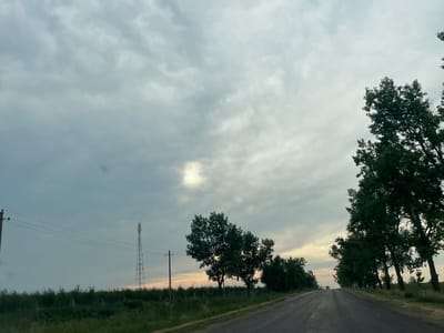 This image depicts a paved rural road in Balabanu, Moldova, as seen from a vehicle. The sky is overcast with heavy, grey clouds, though a patch of brighter sky with sunlight breaking through is visible in the upper left quadrant. A line of tall, green trees runs along the right side of the road, their branches reaching towards the sky. On the left side of the road, a utility pole with multiple wires stretches across the frame, and further in the distance, a tall, lattice-style communication tower stands against the cloudy sky. The foreground shows a grassy verge and a wire fence. The overall impression is of a quiet, overcast day on a country road, possibly late afternoon or early evening, given the subdued lighting. There is no visible text in the image.
