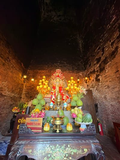 The image depicts the interior of a shrine dedicated to the goddess Thien Y A Na, located in Tuy Hoa Ward, Vietnam. A central statue of the goddess, adorned in red and gold, is the focal point. She is surrounded by offerings, including a variety of fruits such as pomelos, dragon fruit, and watermelons, arranged on a wooden altar. Incense sticks in a dark holder, a golden censer, and decorative yellow flowers also adorn the altar. The shrine's walls are constructed of aged brick, with two vintage-style lamps casting a warm glow. The altar itself is an ornate wooden piece of furniture, decorated with detailed carvings and inlaid mother-of-pearl designs. A red fire extinguisher is visible to the right of the altar. The overall ambiance is one of solemnity and reverence.