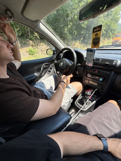 Two individuals are inside a car traveling on a paved road. The driver, a male wearing a brown t-shirt and light-colored shorts, is holding the steering wheel with his left hand. His right arm is raised, and he is touching the car's interior light fixture. A smartwatch is visible on his left wrist. The passenger, also male, is wearing dark shorts and a black t-shirt. The car's dashboard is visible, featuring a navigation system mounted on the windshield, an air freshener hanging from the rearview mirror, a car stereo, and a gear shift. Outside the car, lush green trees and vegetation line the roadside. The road appears to be a rural area, possibly on the outskirts of Daskotna, Bulgaria, given the natural landscape. The lighting suggests it is daytime, with sunlight filtering through the trees.