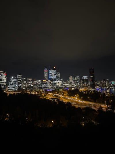The image depicts a nighttime cityscape, with buildings illuminated against a dark sky, likely captured from Kings Park, Australia. The scene includes a stretch of highway with moving vehicles, indicated by the streaks of light. The skyline is composed of numerous tall buildings with lit windows, creating a bright, urban contrast to the surrounding darkness. The right side of the image shows a silhouette of trees, suggesting the edge of a park or green space. The overall environment is urban and modern.