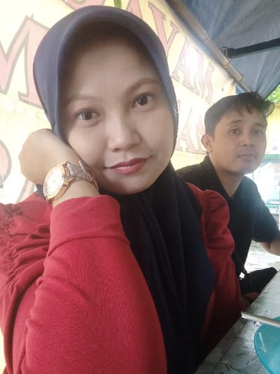 A selfie of a woman wearing a red long-sleeved top and a black hijab. She has a gold watch on her left wrist. A man with dark hair and wearing a black shirt sits behind her to the right. They appear to be at an outdoor restaurant. "AYAM" is visible in large letters on a yellow sign in the background. A blue bowl sits on a metal table in the foreground.