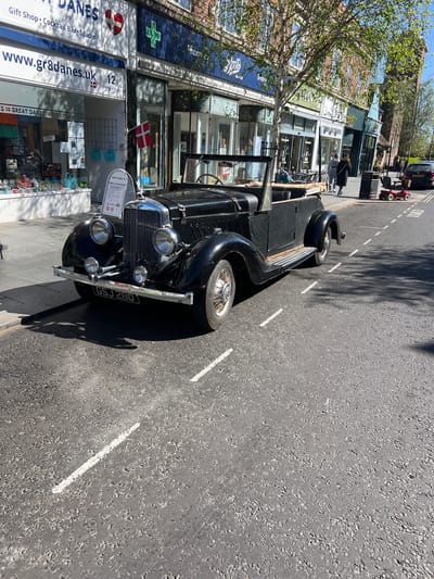 A sunlit street scene in Frinton-on-Sea, United Kingdom, captures a vintage black convertible car parked on a paved street. The classic vehicle, featuring a chrome grille, circular headlights, and wire-spoke wheels, displays the license plate "GSJ 280". Its dark shadow extends across the light-colored pavement, suggesting bright sunshine and clear weather, likely mid-morning or afternoon.

On the left, a row of commercial buildings lines the street. The most prominent establishment is "GREAT DANES," identified as a "Gift Shop • Cocktail Bar • Studio." This shop showcases Danish themes, with a red and white Danish flag flying and a heart symbol incorporating the Danish cross. Its website, "www.gr8danes.uk," and address, "12 Connaught Ave," are visible on the storefront. Other shop windows reveal merchandise and an "OPEN" sign.

Two people are walking on the sidewalk in front of the shops. Further down the street, a small red toy car or scooter is visible near the curb. The asphalt road features white dashed markings. Trees with fresh green leaves partially obscure the upper left portion of the buildings, adding a touch of natural vibrancy to the urban setting.