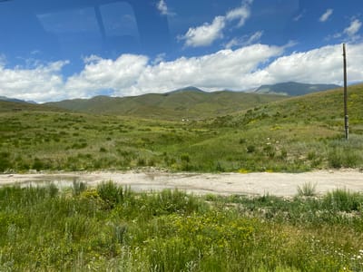The image depicts a vast, rolling green landscape under a bright blue sky dotted with fluffy white clouds. The terrain is characterized by undulating hills covered in lush grass and scattered wildflowers in yellow and white hues. A dirt road runs horizontally across the foreground, with a large puddle of water reflecting the sky. Beyond the road, the landscape stretches towards distant mountains, some of which are shrouded in atmospheric haze. A single wooden utility pole stands on the right side of the frame, with power lines stretching across the middle ground. In the far distance, small structures, possibly buildings or tents, are visible nestled within the rolling hills. The overall impression is one of a serene, natural, and remote environment. The image is taken from an elevated perspective, possibly from a vehicle, as indicated by a subtle reflection on the top left portion of the image. The location is identified as being in the vicinity of Kaysar, Kyrgyzstan.
