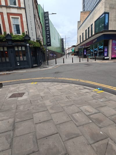 The image shows a street scene in Newport, United Kingdom, likely near the city center. The foreground is dominated by a paved sidewalk with large, square grey paving stones. A yellow painted marking and a discarded plastic water bottle are visible on the sidewalk. The street itself is asphalt with double yellow lines painted on it, indicating parking restrictions.

Buildings line the street. On the left is a pub with a dark facade and large windows, adorned with hanging plants and signage. Behind and to the right of the pub, a modern building with a green facade is visible, featuring large, vertically oriented "DEBENHAMS" banners. Further down the street, a concrete building with large windows and a blue trim is visible, appearing to be a retail or entertainment establishment with digital screens displaying advertisements, including one for "HELLO NEWPORT." A street sign reading "CARDIFF STREET" is partially visible.

The sky is overcast and grey. The scene appears to be unoccupied by any people, suggesting it was taken at a time when the street was not busy. The overall atmosphere is one of an urban environment, possibly during off-peak hours.