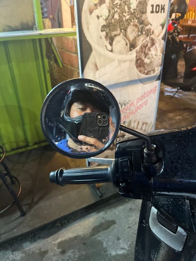 A selfie is reflected in the side mirror of a dark-colored motorcycle parked on a wet street in Boja, Indonesia. The rider, wearing a black helmet and a blue shirt, holds a smartphone in their right hand to take the photo. The smartphone has a black case with a prominent camera bump. In the background, a poster with food images and text, including "10K" and "eam potong singapore," is visible. The street is dimly lit, suggesting it is nighttime or early morning, and water droplets are scattered across the motorcycle's surface and the ground. A green-colored structure, possibly a stall or building facade, is also present in the background to the left.