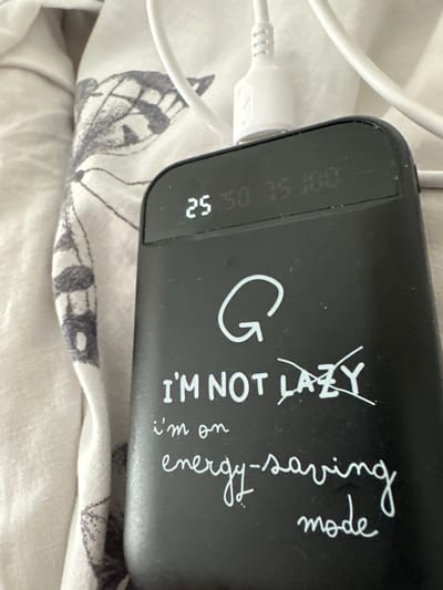 The image shows a close-up view of a black power bank with white text. The power bank is resting on a white and grey patterned fabric, likely a bedspread. A white charging cable is plugged into the top of the power bank. The power bank has a digital display on the top that shows "25 50 75 100". The text on the power bank reads "G I'M NOT LAZY i'm on energy-saving mode". The overall scene is domestic and casual. There is no indication of the city of Dolni Chiflik, Bulgaria in the image.